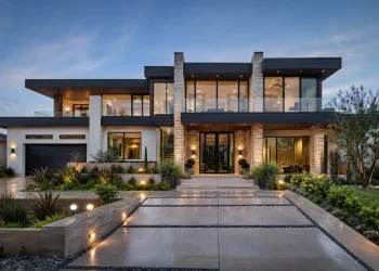 Front exterior of a modern luxury home at dusk, featuring a flat roof, large glass windows, stone pillars, and illuminated landscaping.