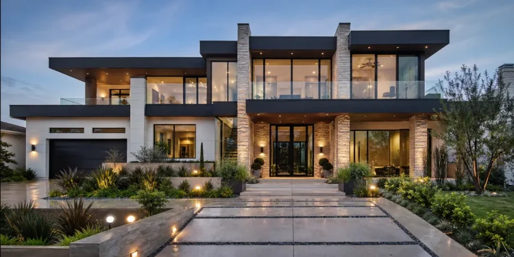 Front exterior of a modern luxury home at dusk, featuring a flat roof, large glass windows, stone pillars, and illuminated landscaping.