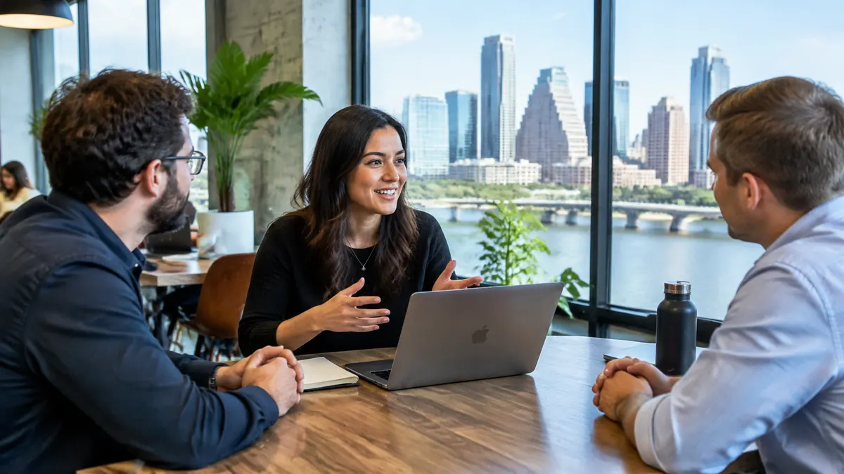 Three professionals discuss work around a laptop in a city-view office.