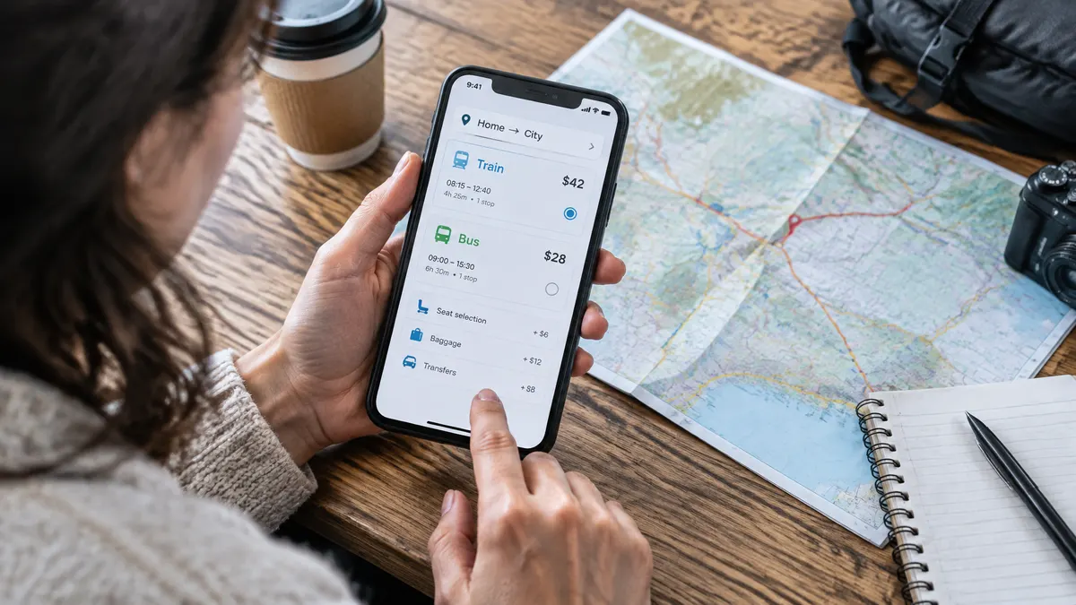 Traveler compares train and bus fares on a phone beside a map and travel-planning supplies.